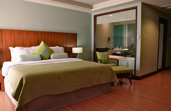 Una habitación de hotel con una cama grande, cabecera de madera, una zona de estar con una silla y una mesita pequeña, y un baño visible a través de una puerta abierta.
