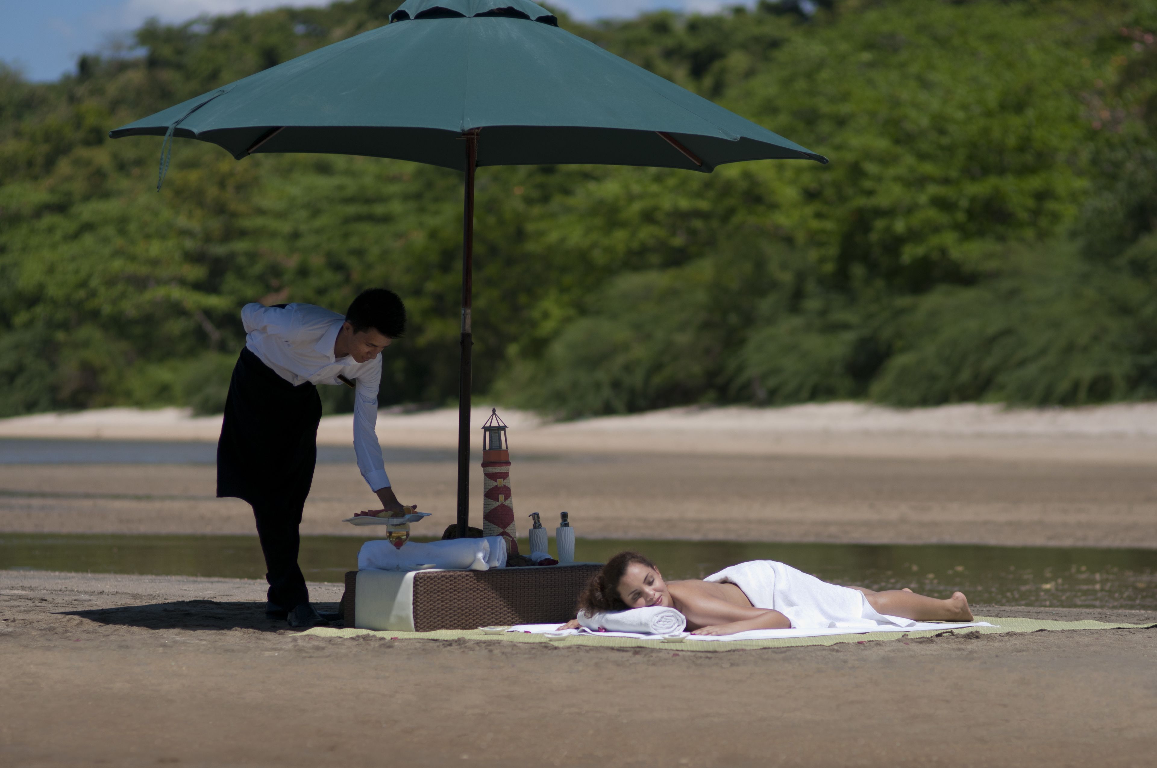 A person lying on a towel under a green umbrella on a sandy beach with a water body and greenery in the background.