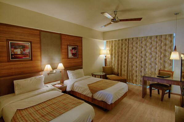 A hotel room with two beds, a desk, a chair, a ceiling fan, and a large window with curtains.