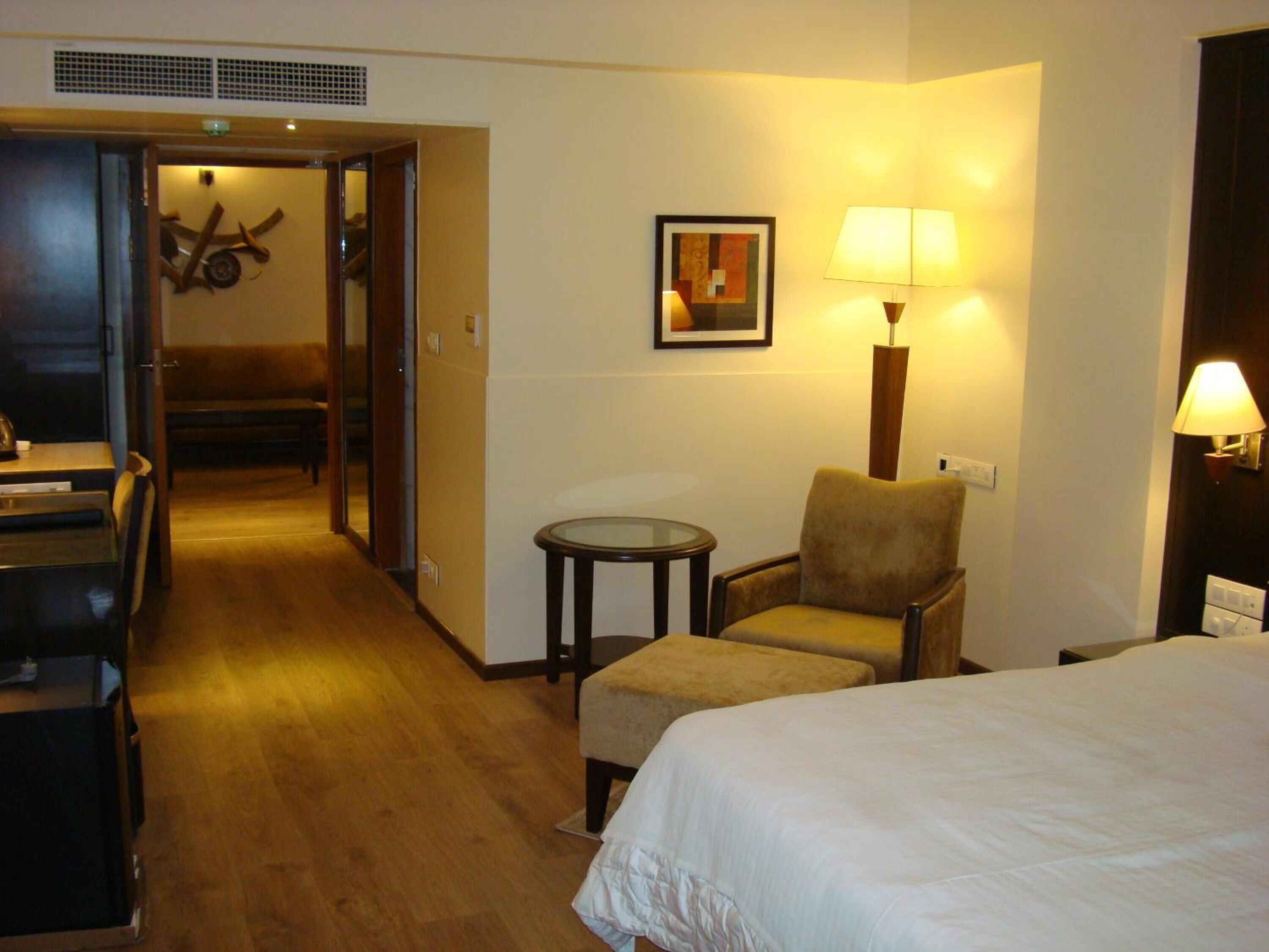 Habitación de hotel con cama, una silla, una mesita y una lámpara. Se ve una puerta que da a un pasillo con un banco y un adorno en la pared.