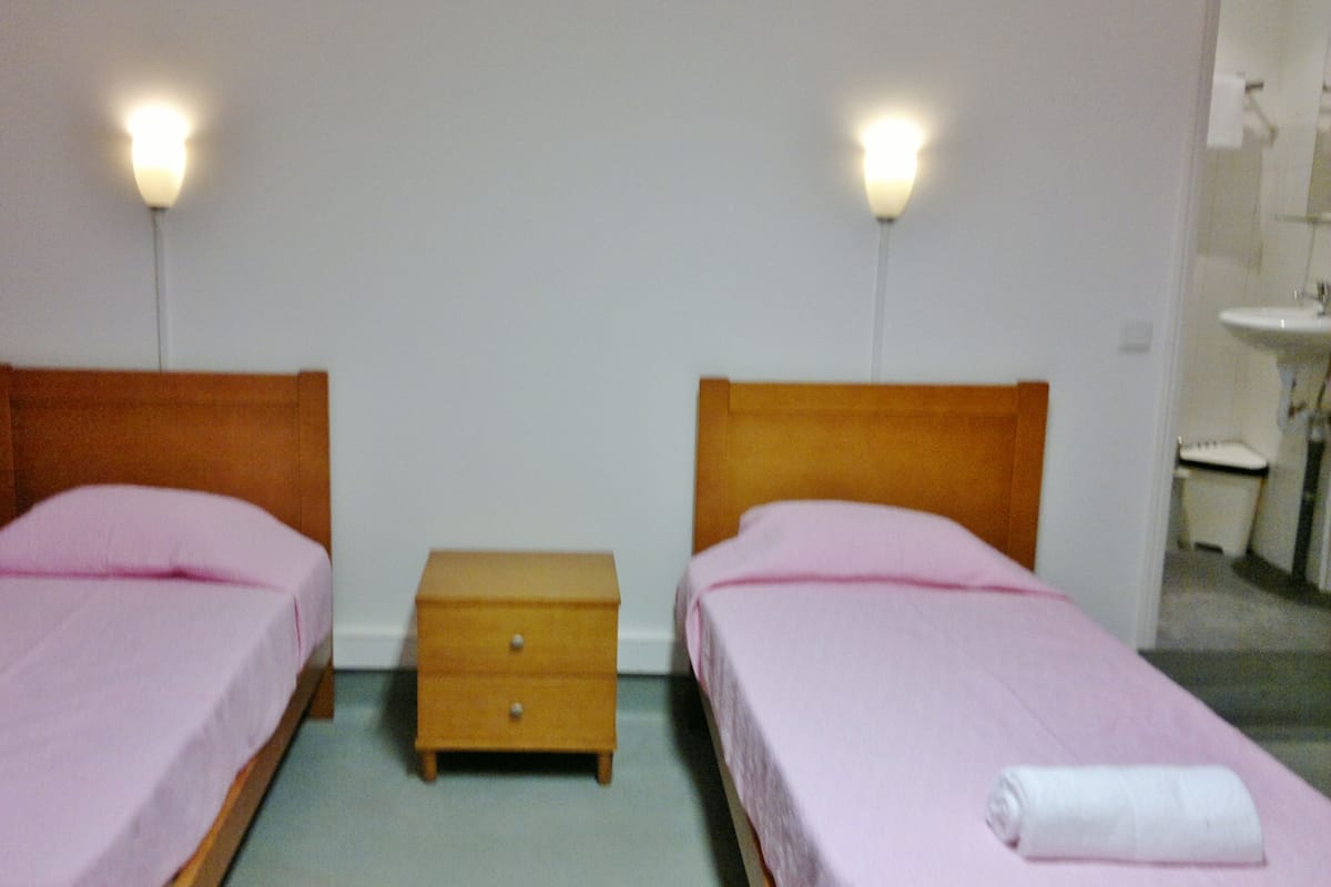 A twin room with two beds, each with a wooden headboard and pink bedding. There is a small wooden nightstand between the beds with a drawer.