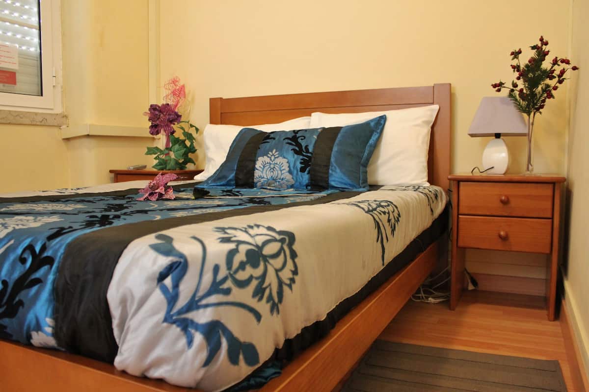 A neatly made bed with a floral bedspread, a bedside table with a lamp, and a vase with flowers.