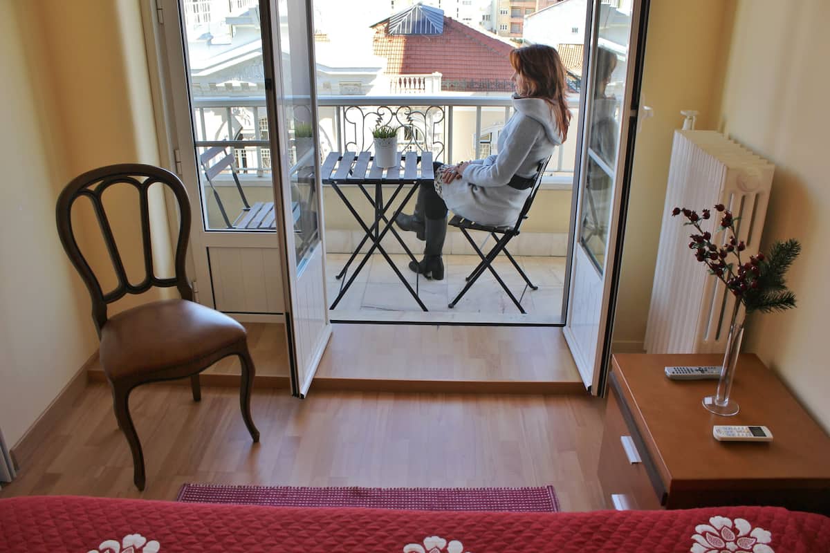 A room with a balcony, a chair, a small table with a potted plant, a radiator, and a vase with flowers.