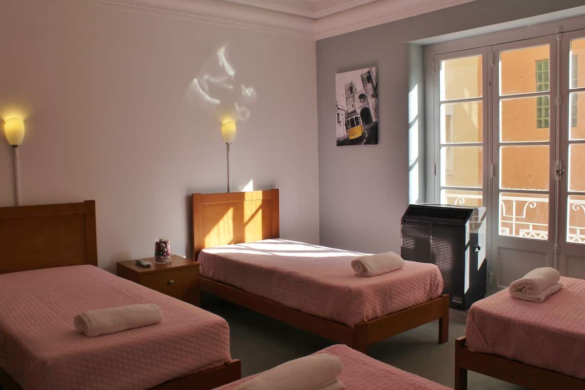 A hotel room with two single beds, a nightstand, a lamp, a radiator, and a window with a view of buildings.