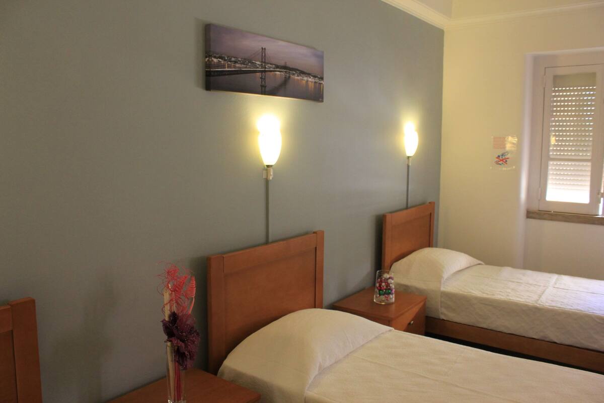 A hotel room with two single beds, a nightstand with a lamp, a decorative vase, and a painting of a bridge on the wall.