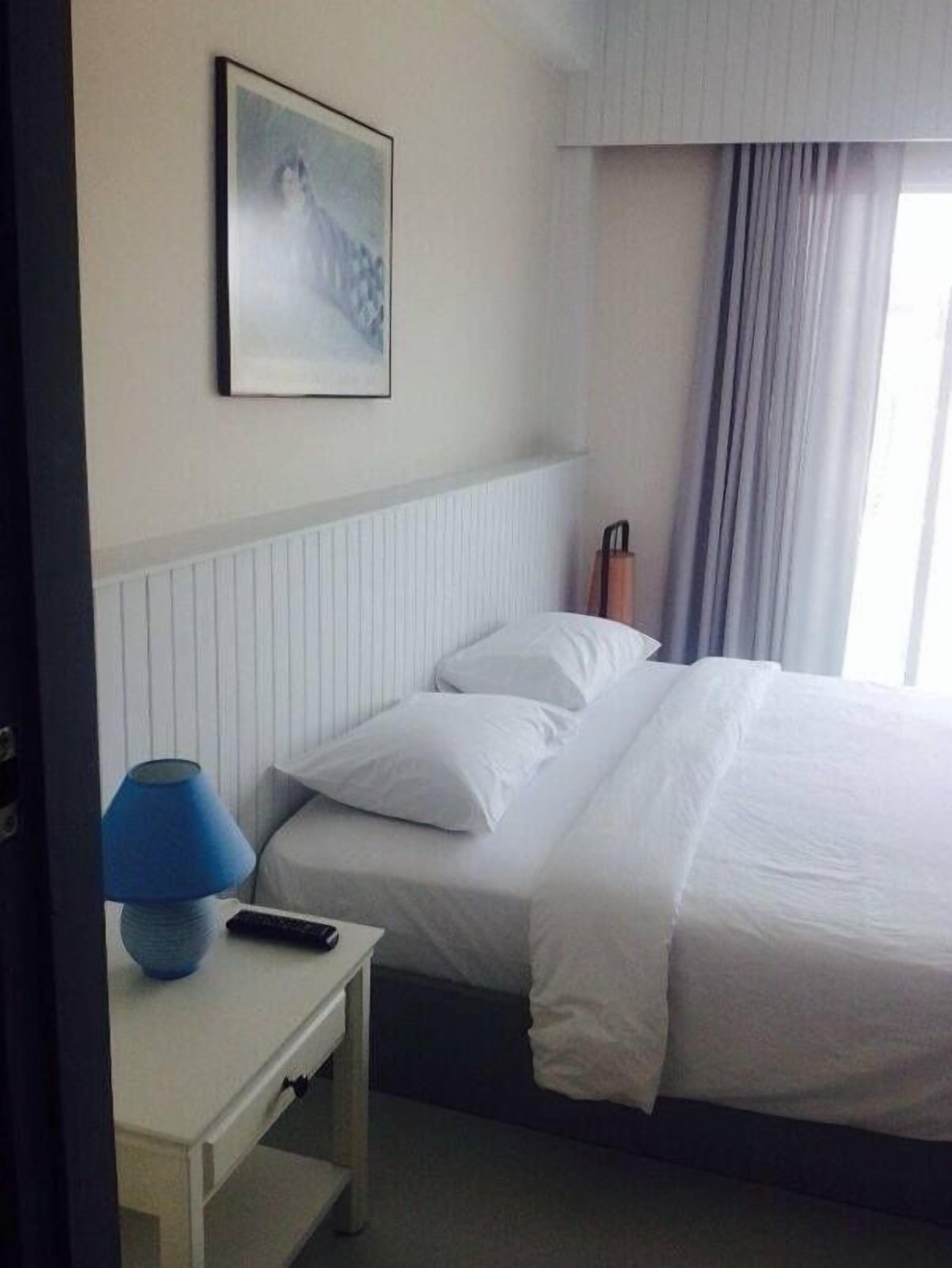 A hotel room with a bed, a nightstand with a lamp, a painting on the wall, and a window with curtains.