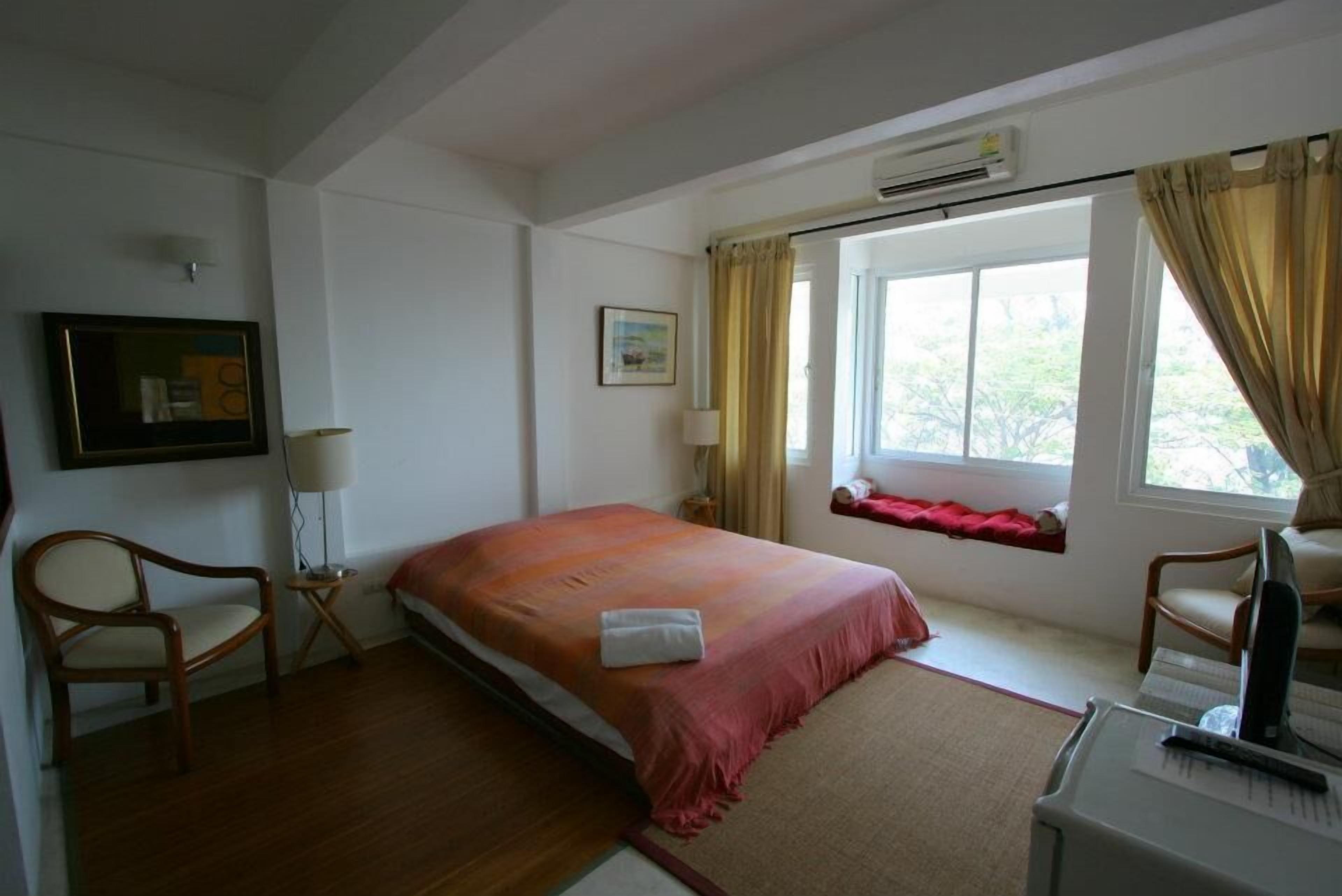Un dormitorio con cama, dos sillas, un escritorio con computadora, una ventana con cortinas y un cuadro en la pared.