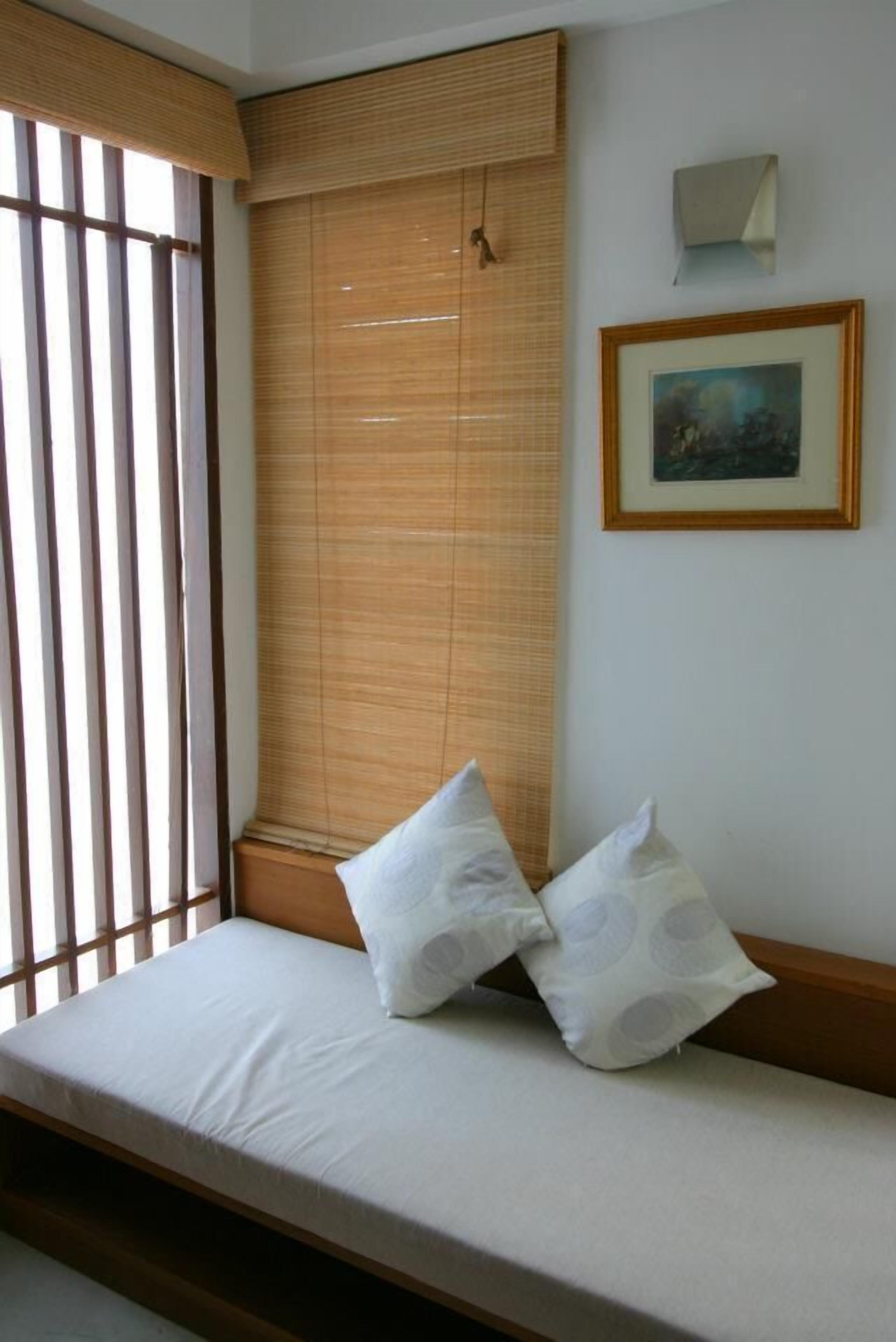 A room with a bed, a window with vertical blinds, a framed picture on the wall, and a small wall-mounted light.