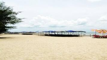 Una playa con hileras de sombrillas y sillas de descanso, un muelle que se adentra en el agua, y un cielo despejado.