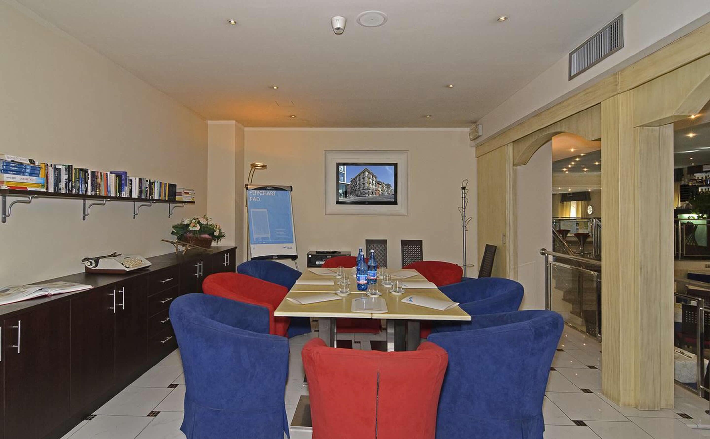 A modern dining area with a long table, blue and red chairs, a bookshelf with books, a framed picture on the wall, and a view of the outside through a glass door.