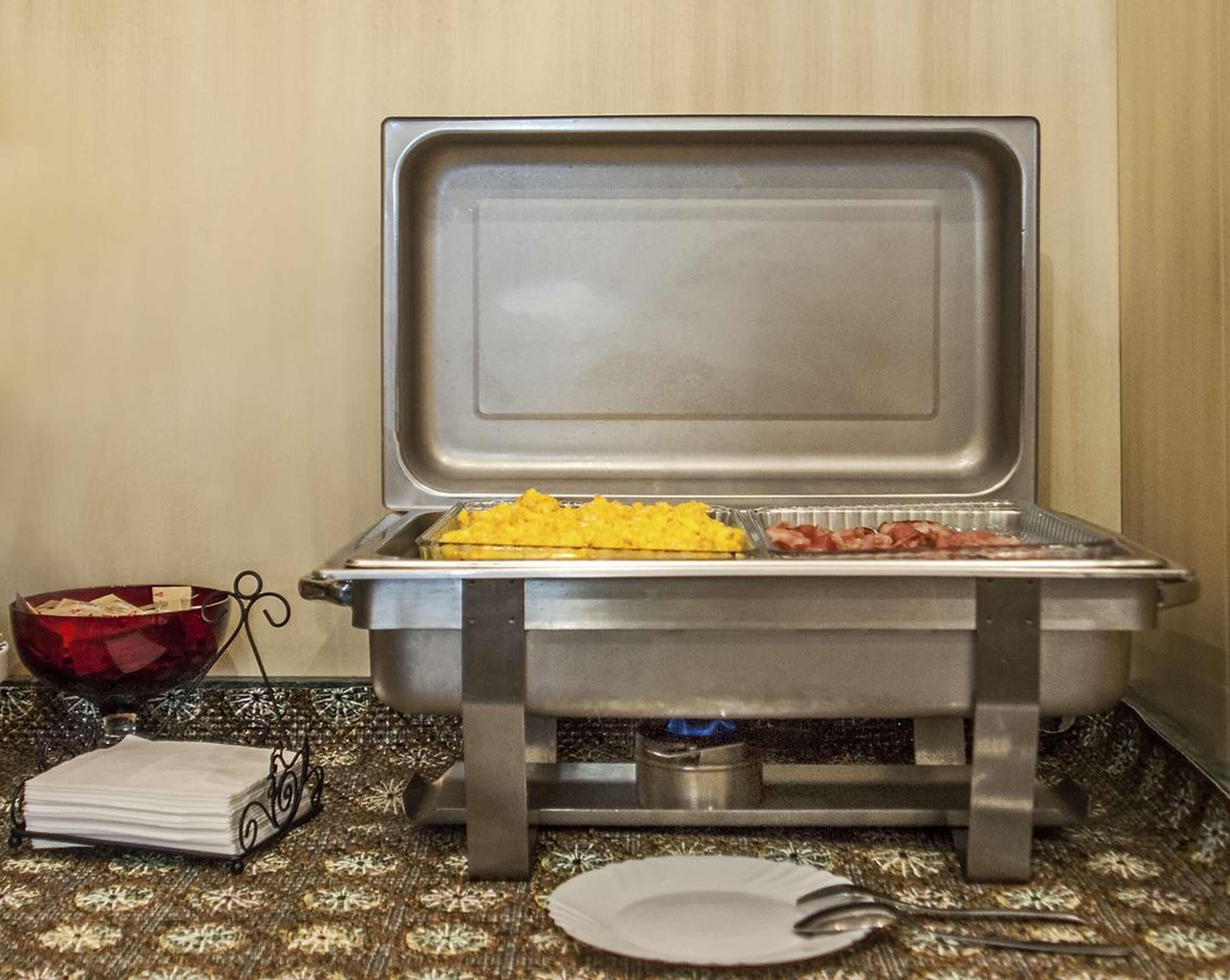 A buffet setup with a tray of scrambled eggs and bacon, a bowl of fruit, and a napkin holder on a patterned tablecloth.