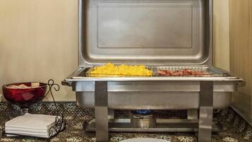 A buffet setup with a tray of scrambled eggs and bacon, a bowl of fruit, and a napkin holder on a patterned tablecloth.
