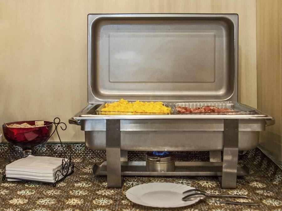 A buffet setup with a tray of scrambled eggs and bacon, a bowl of fruit, and a napkin holder on a patterned tablecloth.