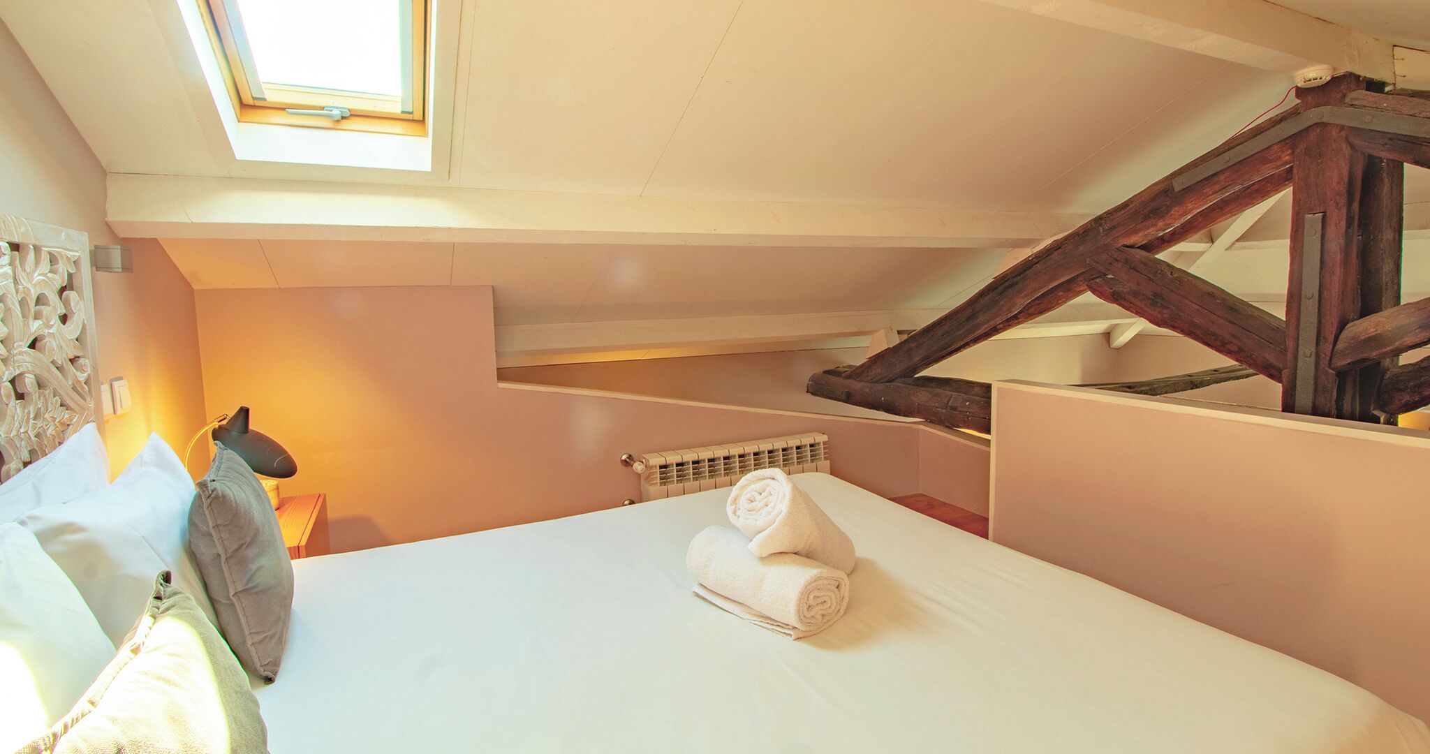 Quarto com claraboia, cama com toalhas dobradas, radiador e estrutura de vigas de madeira.