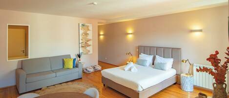 A modern bedroom with a grey sofa, a large bed with white linens, a small round table, and a decorative vase.