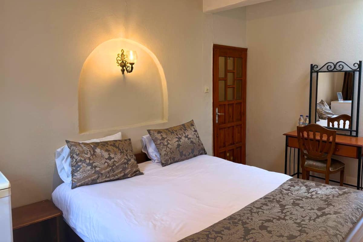 A hotel room with a bed, a desk with a chair, a mirror, and a wall-mounted light fixture.