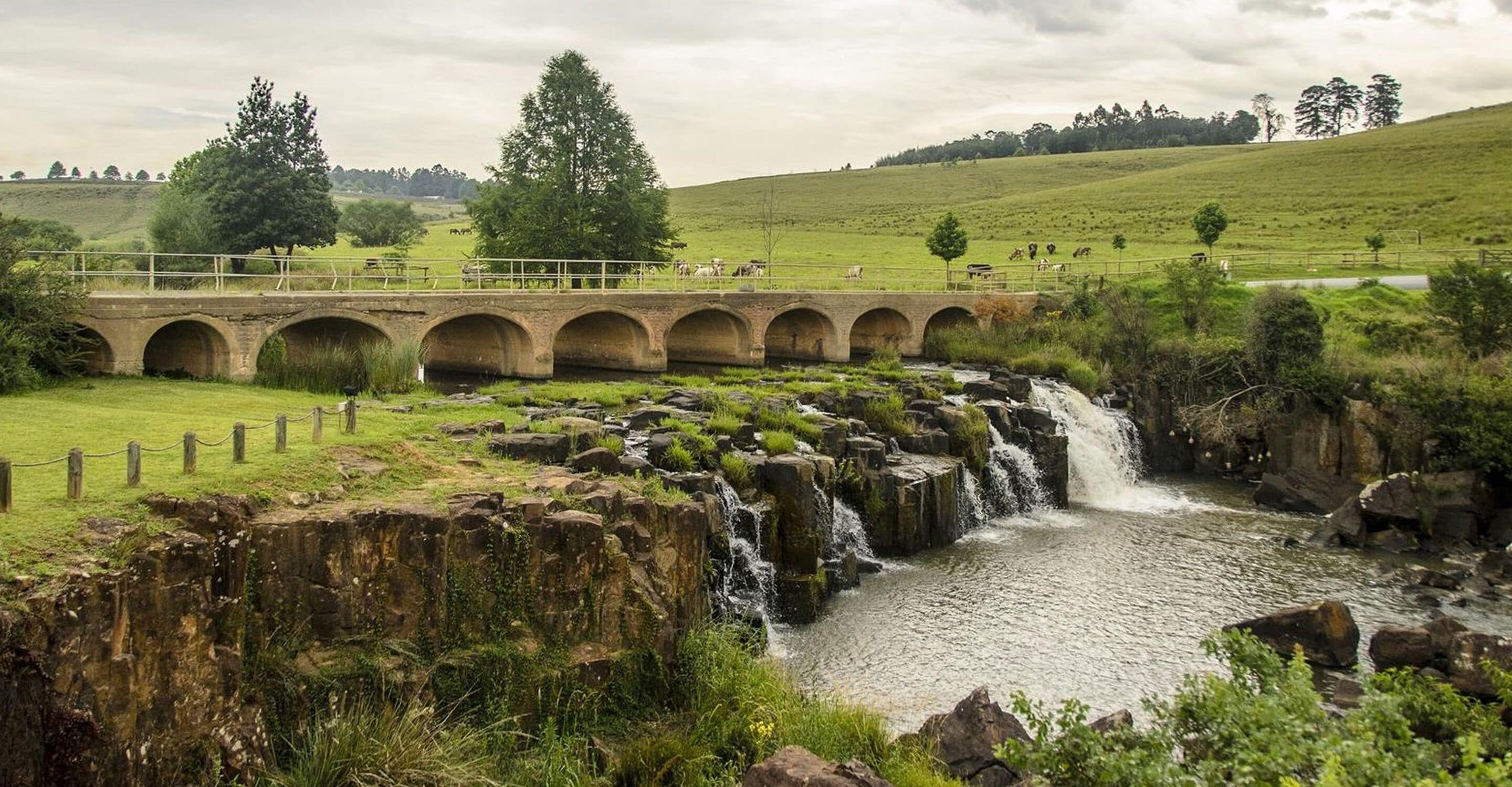 Un puente de piedra sobre un río con una cascada, rodeado de campos verdes y árboles.