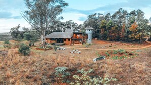 A rustic cabin with a large deck, surrounded by dry grassland and trees, featuring a water tower and a small tower structure.