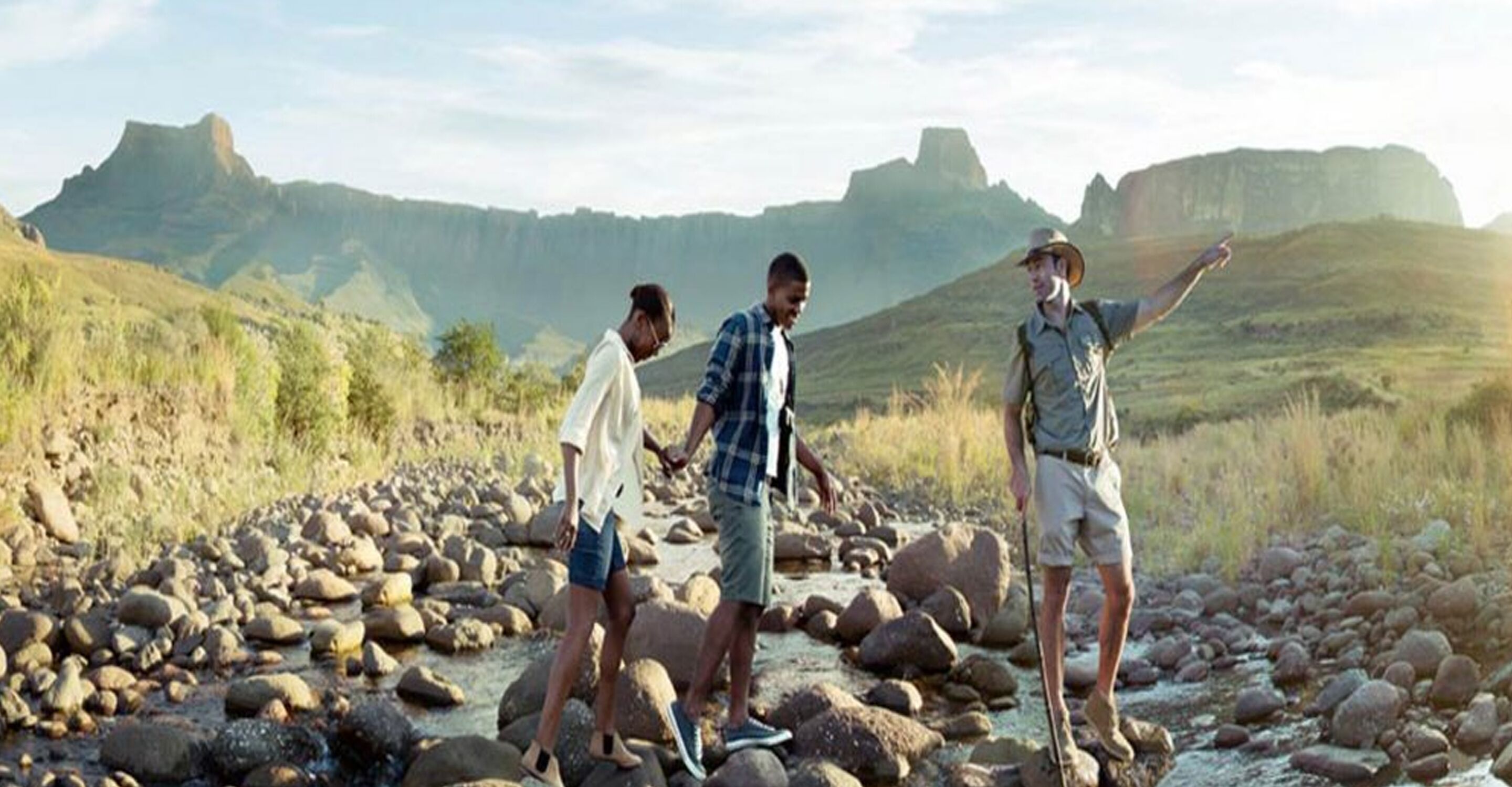Three people standing on a rocky riverbed with mountains in the background.