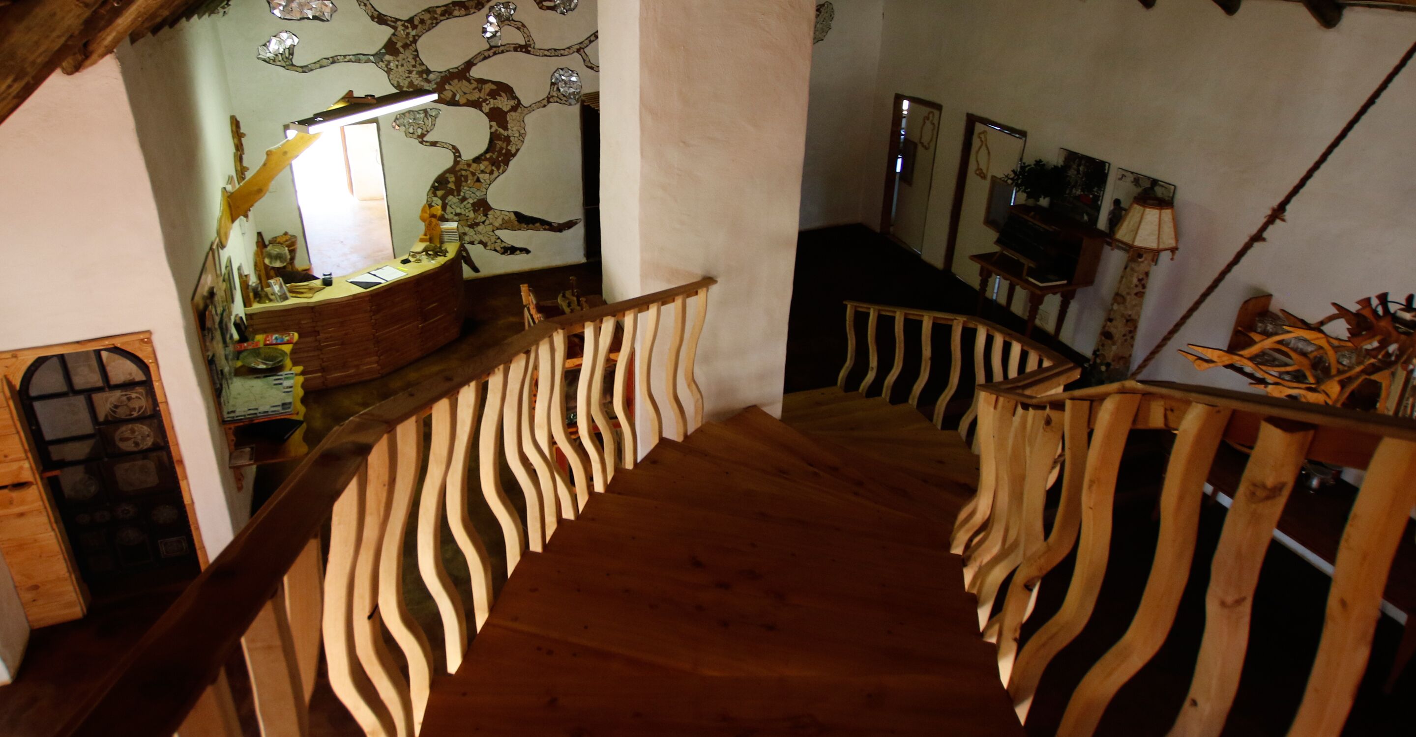 Un escalier en bois avec une rampe ondulée menant à une pièce ornée d’une fresque représentant un arbre, où se trouvent un bureau et une lampe.