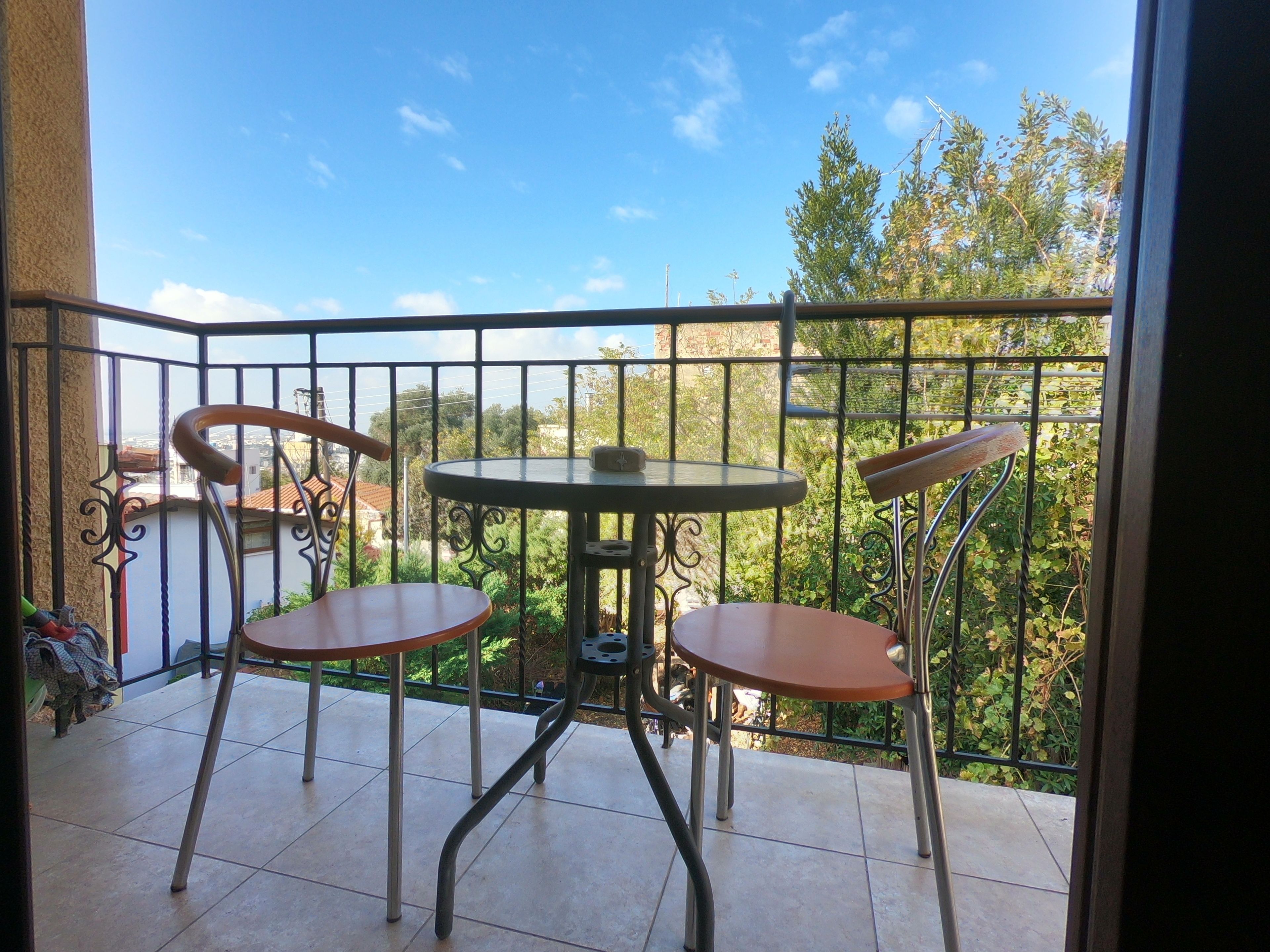 A balcony with a round table and two chairs, a view of greenery and buildings in the background.