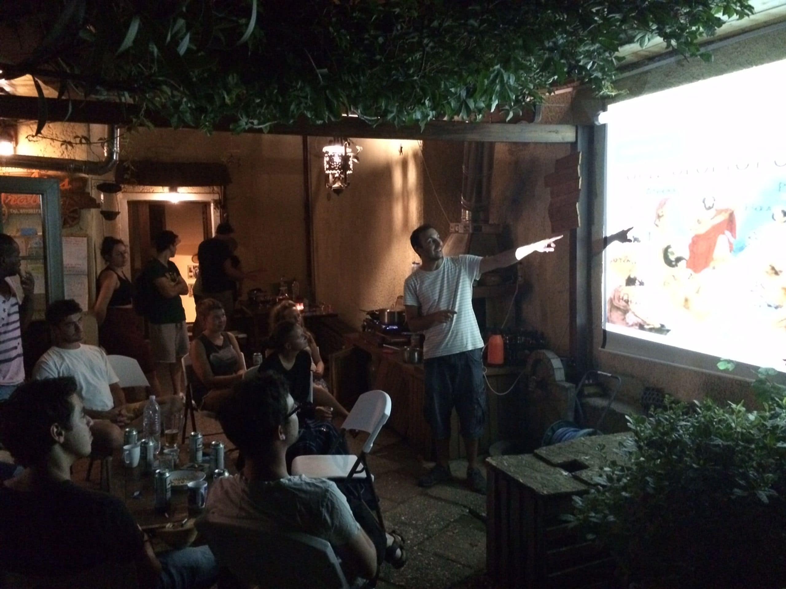 A group of people gathered around a projector screen in an outdoor setting with tables and chairs.