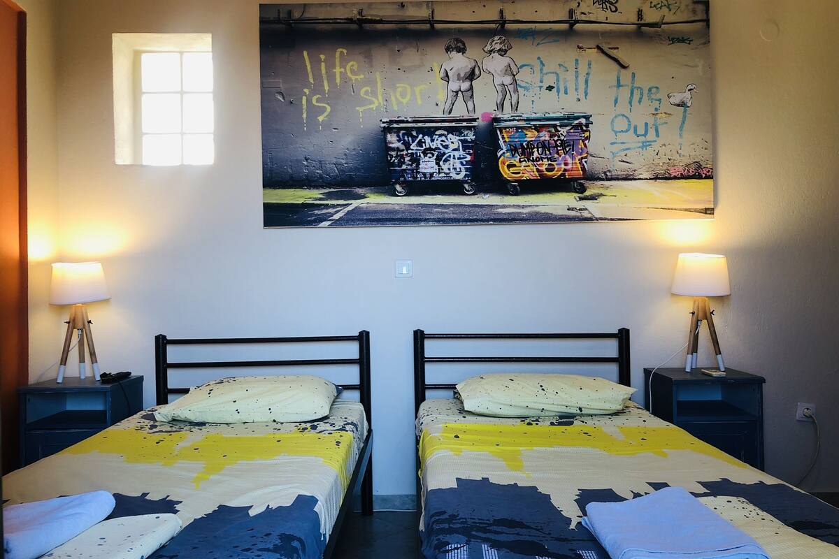 A room with two beds, each with a patterned bedspread and pillows. There are two bedside tables with lamps and a wall-mounted painting above the beds.