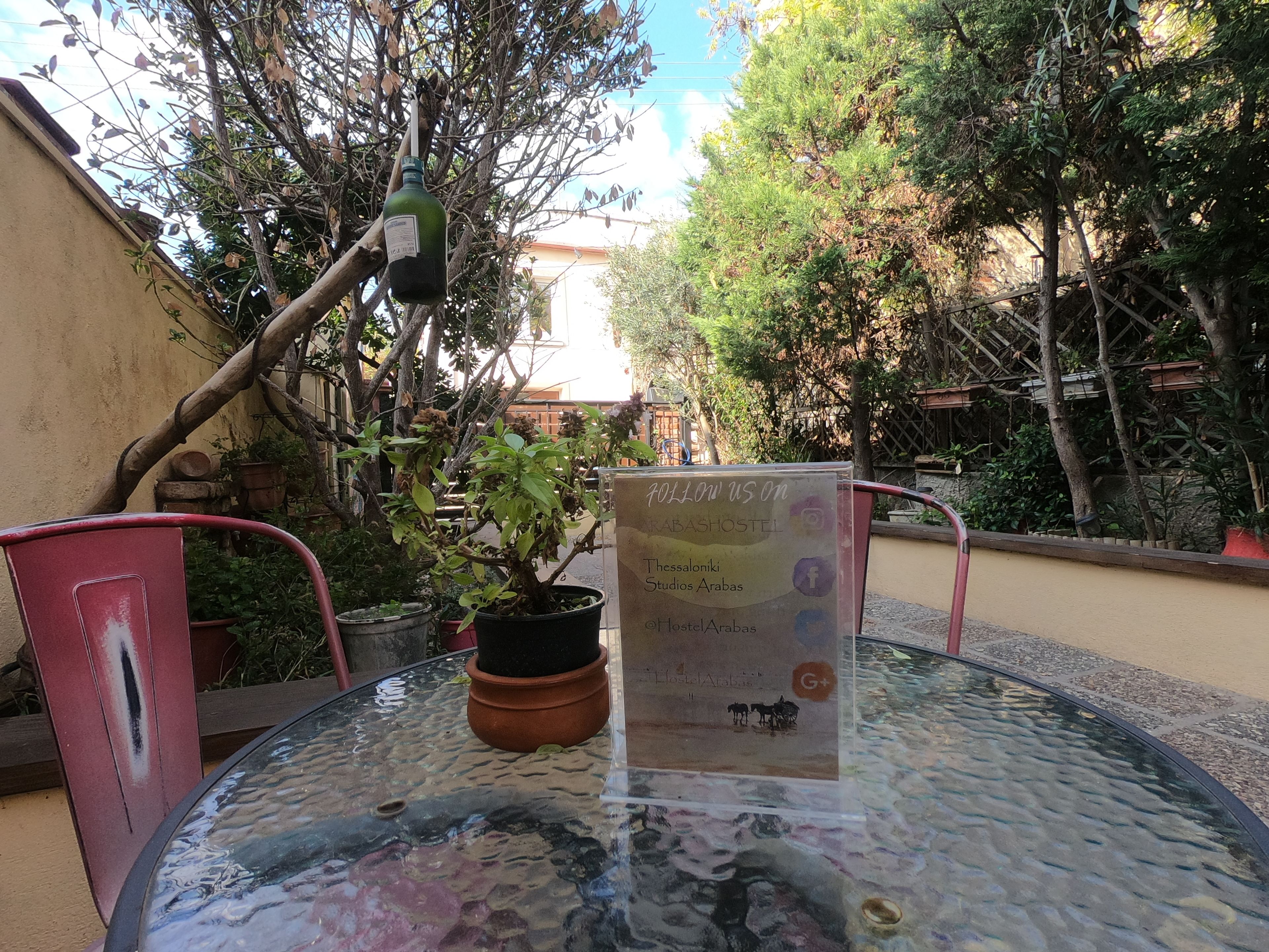 A glass table with a sign for 'HostArabas' in a courtyard with potted plants and a hanging bottle.