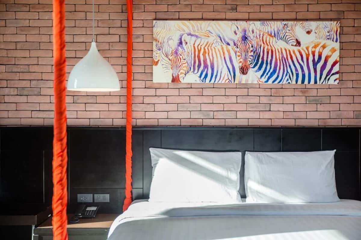 Hotellihuone, jossa on tiiliseinä, seepramaalauksia, valkoinen sänky ja katosta roikkuva valaisin.