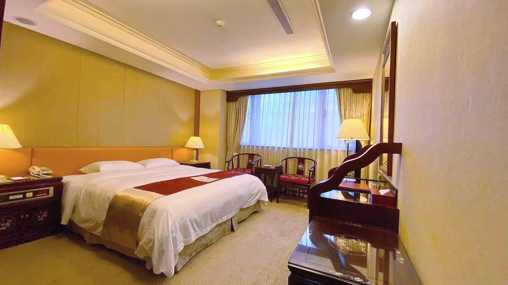 A hotel room with a large bed, two bedside tables with lamps, a desk with a chair, a mirror, and a window with curtains.