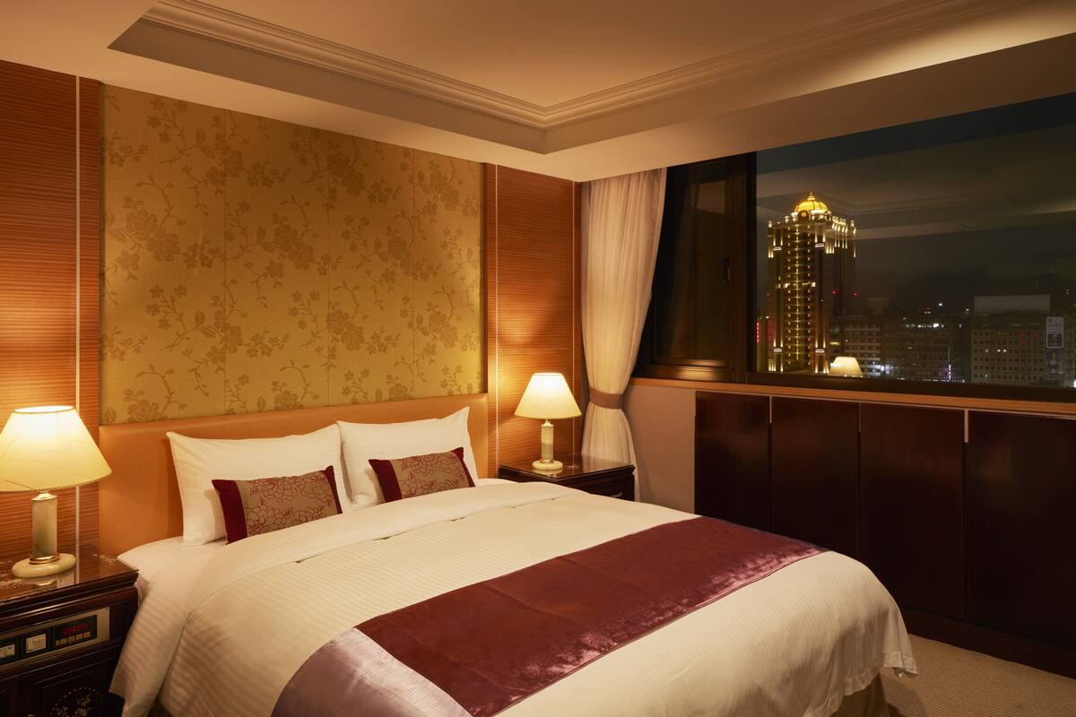 Ein Hotelzimmer mit einem großen Bett, zwei Nachttischen mit Lampen, Blick auf eine Stadtlandschaft durch ein Fenster und einem dekorativen Kopfteil.