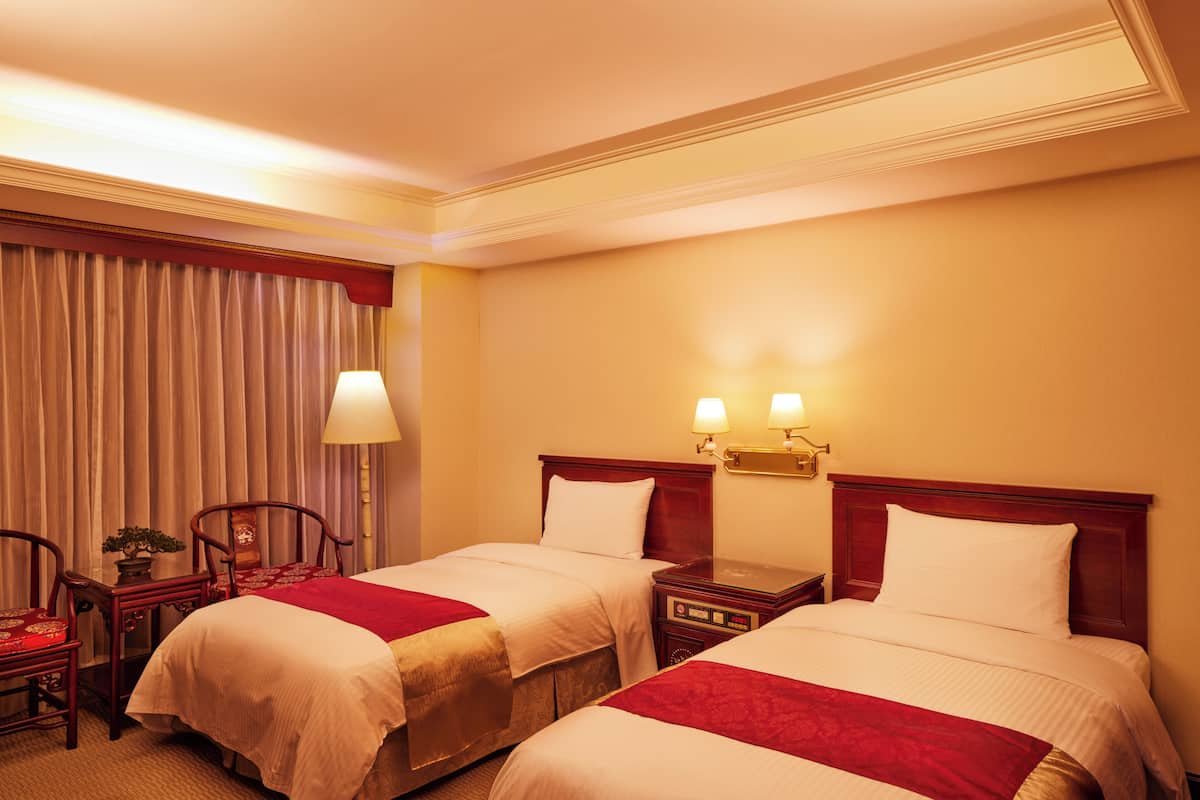 Ein Hotelzimmer mit zwei Betten, einem Schreibtisch mit Lampe, einem Sessel und einem kleinen Tisch mit Pflanze.