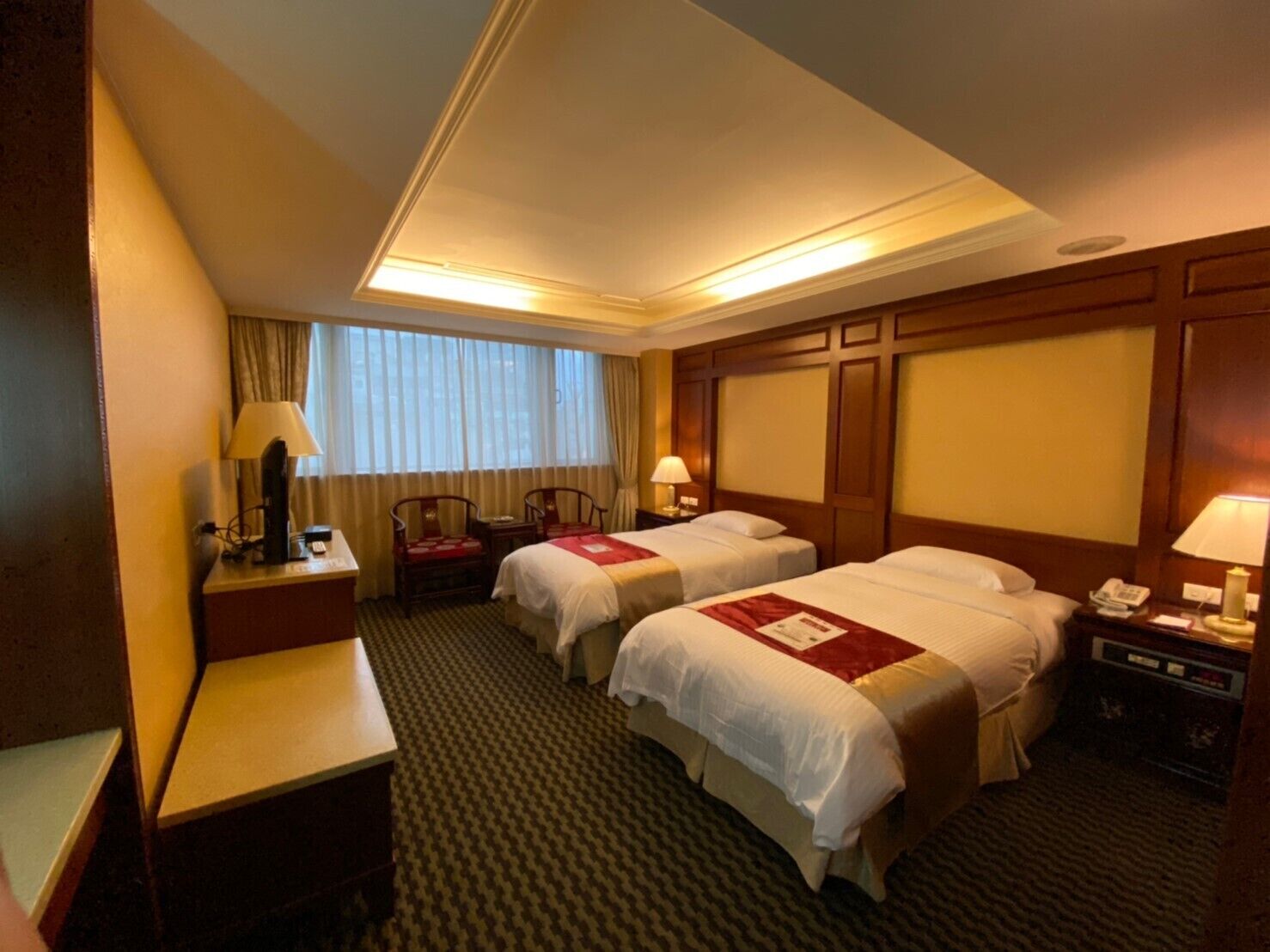 A hotel room with two beds, a desk, a chair, a window with curtains, and a nightstand with a lamp.