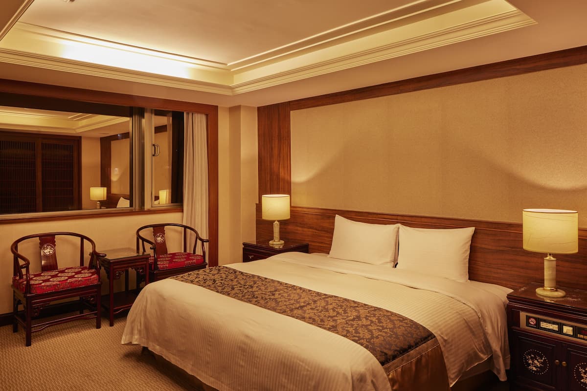 Ein Hotelzimmer mit einem großen Bett, zwei Nachttischen mit Lampen, einem Holz-Kopfteil, einem Spiegel und zwei Polsterstühlen.