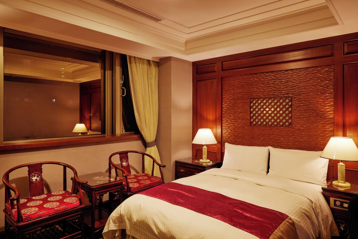 Ein Hotelzimmer mit einem großen Bett, zwei Nachttischen inklusive Lampen, einem Holz-Kopfteil, einem Fenster mit Vorhängen und zwei Holzstühlen mit roten Kissen.