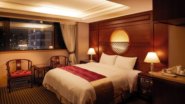 Habitació d'hotel amb un llit, dues taules de nit, una finestra amb cortines i una vista nocturna de la ciutat.