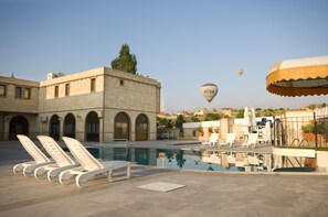 A pool area with lounge chairs, a hot air balloon in the distance, and a building with arched windows. - Ciner Hotel (Nevsehir)