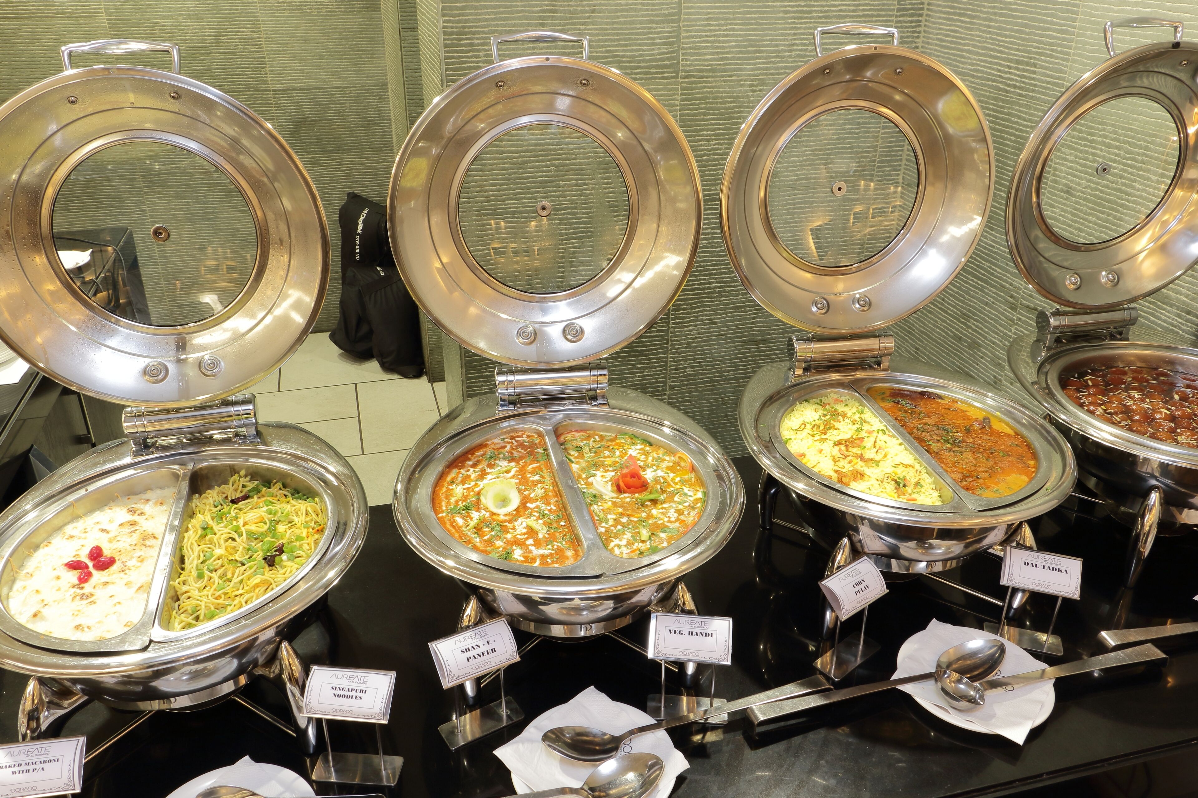 A buffet with multiple food stations, each with a metal lid and labeled with different dishes.