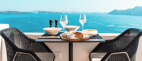 Una mesa dispuesta con un plato de comida, dos copas de vino blanco y saleros y pimenteros en un balcón con vistas al agua.
