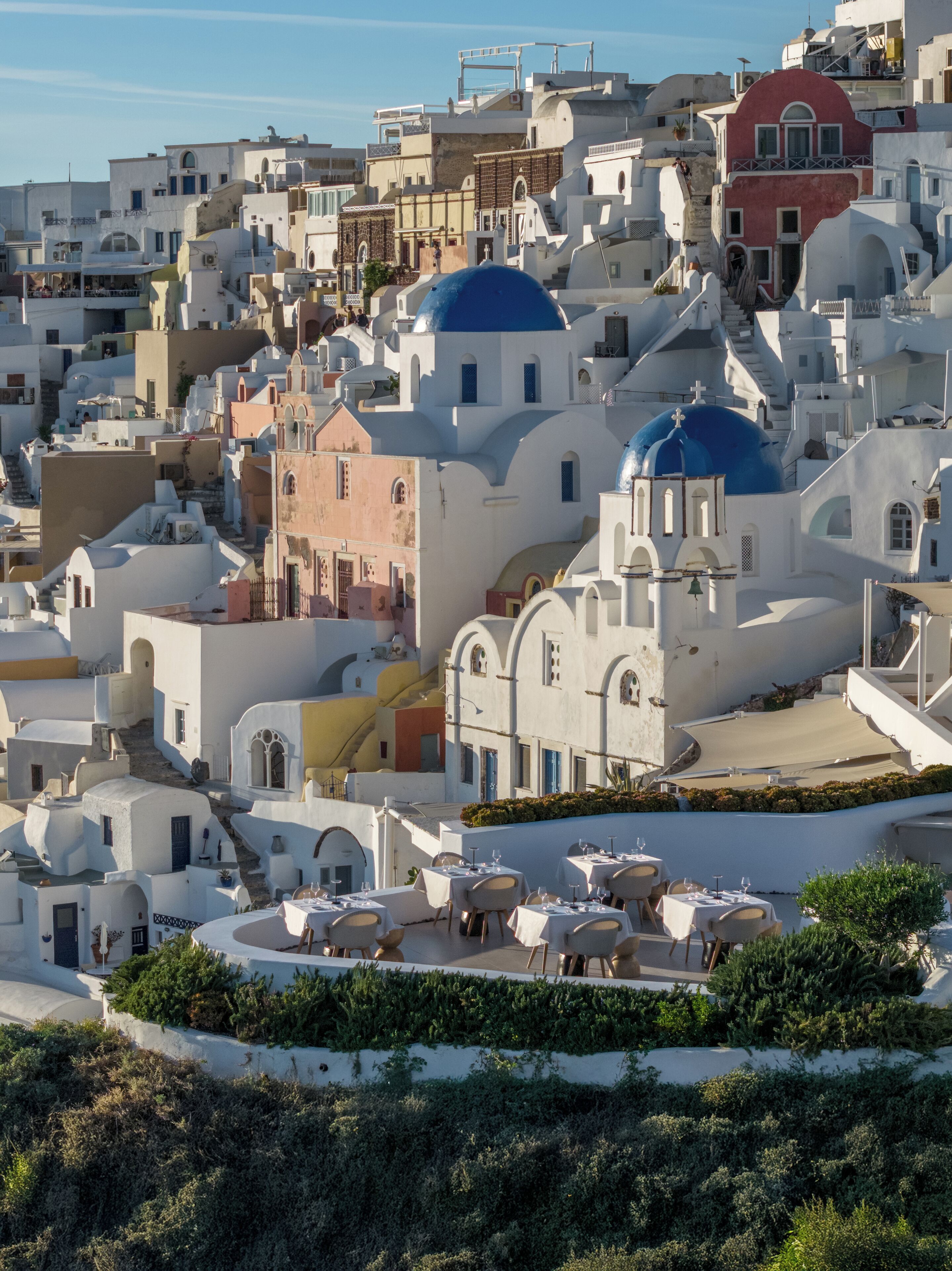 Un pueblo costero con edificios blancos y cúpulas azules, con terrazas al aire libre y una estructura en pendiente.