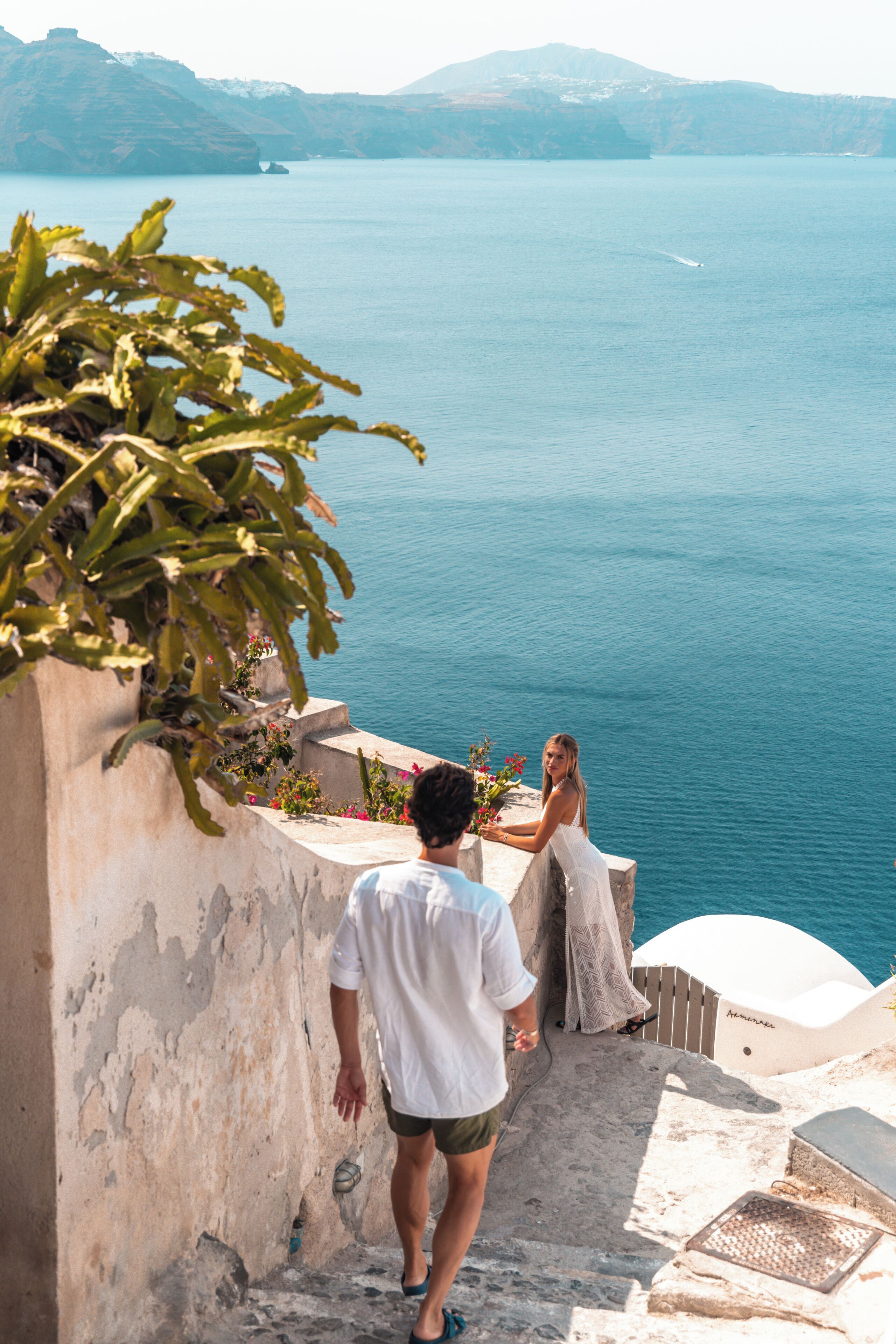 Um homem e uma mulher em uma escadaria de pedra, com vista para um corpo d'água e montanhas ao fundo.