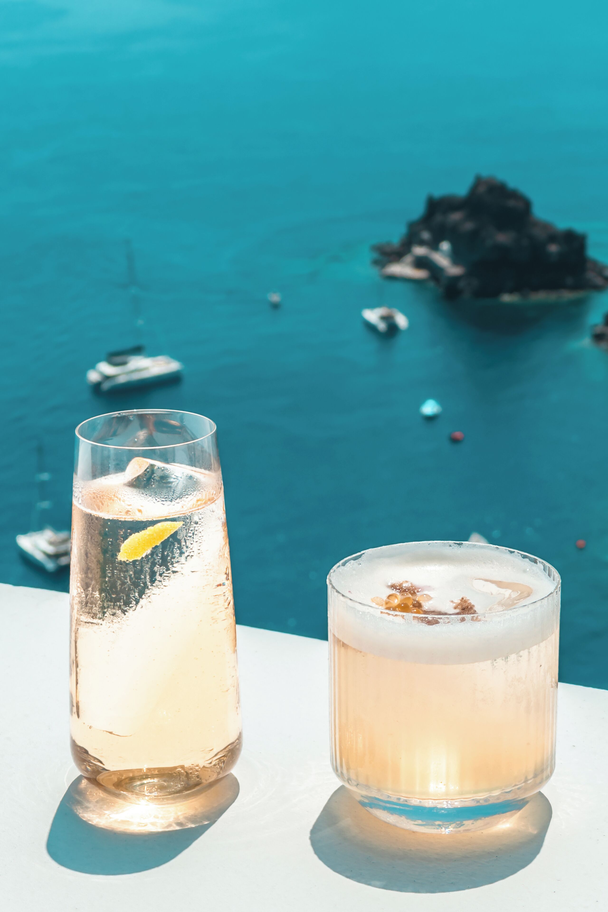 Dos vasos de bebidas sobre una superficie blanca, con vista a un cuerpo de agua y barcos al fondo.