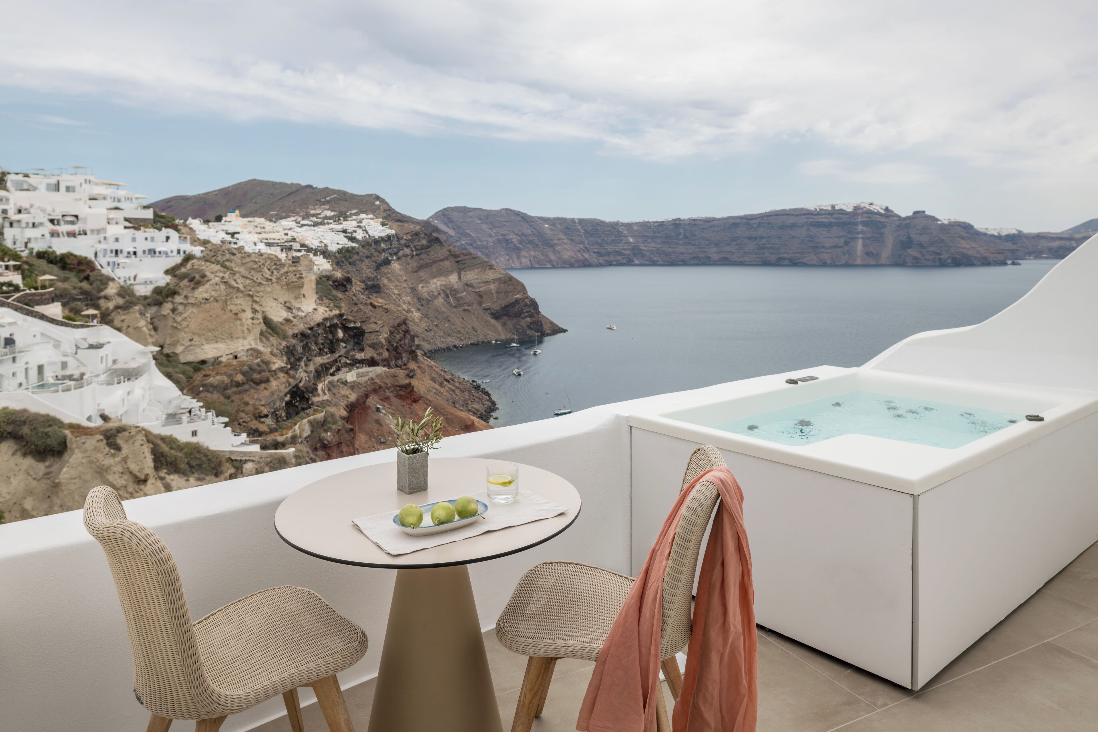 Un balcón con una bañera blanca, sillas de mimbre, una pequeña mesa con frutas y vistas a un paisaje costero con edificios blancos.
