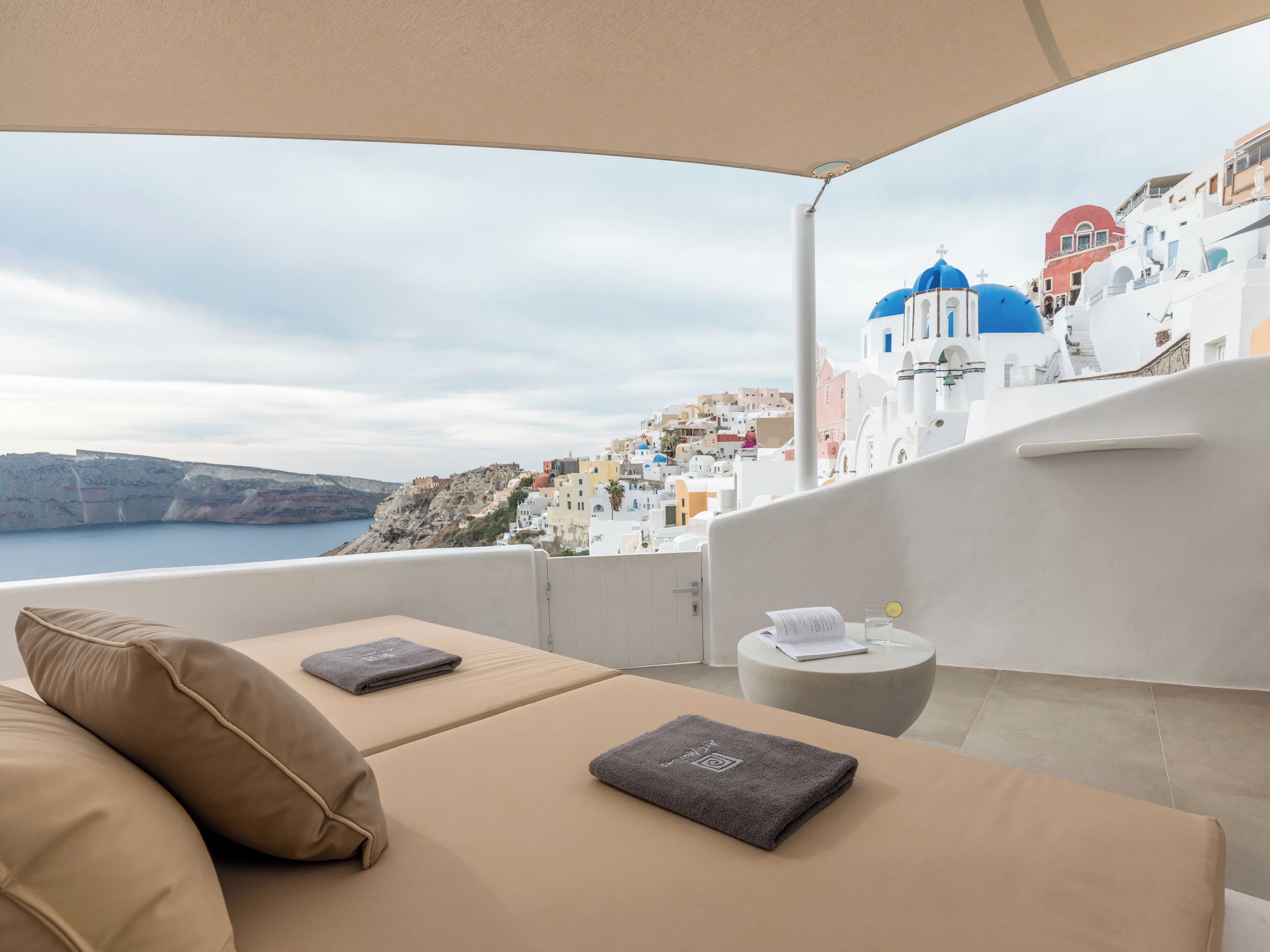 A balcony with a view of a coastal town, a sofa, and a small table with a book and a glass.