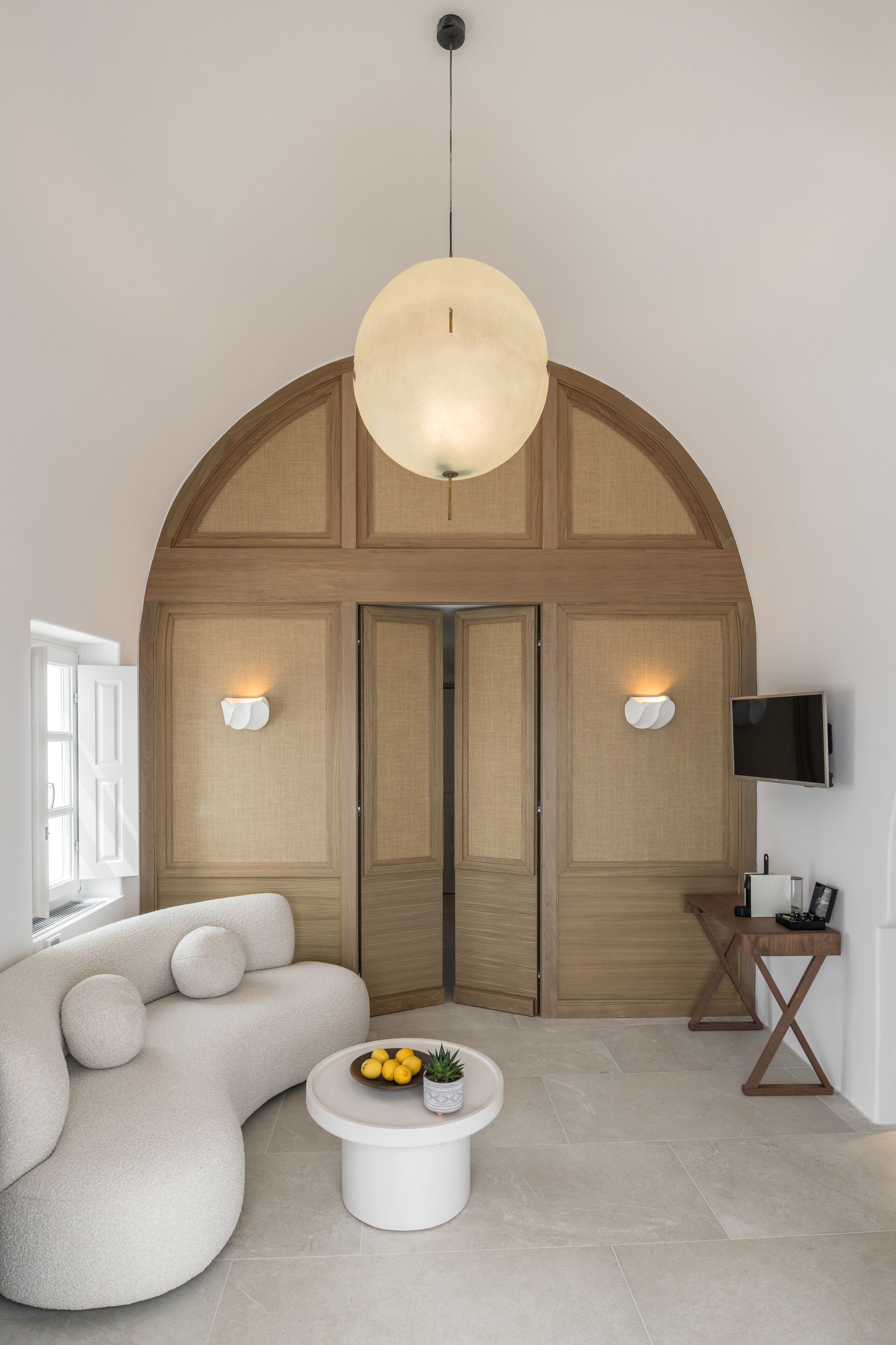 Un salon moderne doté d’une porte en bois à arc unique, d’une table basse ronde blanche agrémentée d’une plante et d’un canapé aux lignes épurées.