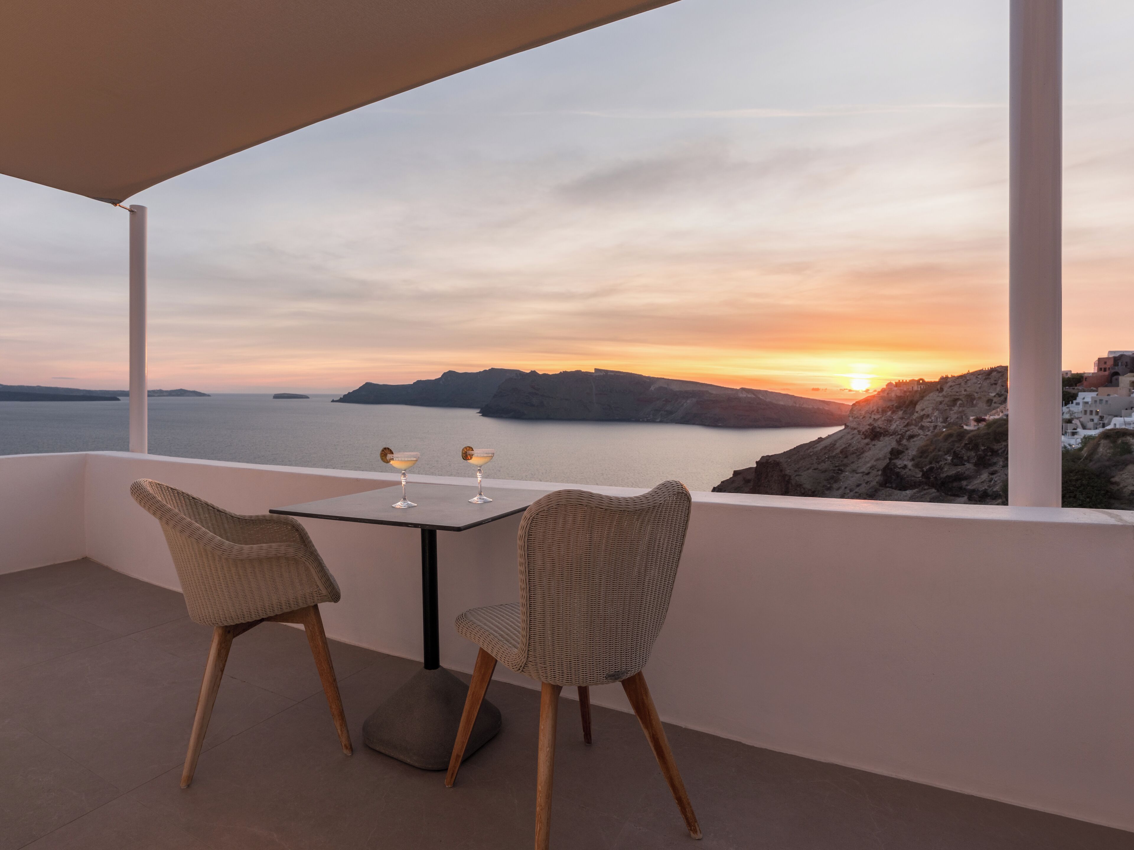 Un balcón con dos sillones de mimbre y una mesa, con vistas a una puesta de sol en la costa.