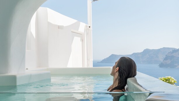 Una mujer se relaja en una piscina infinita transparente con vistas a una costa montañosa.
