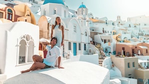 Una pareja sentada sobre un borde de piedra blanca, con vista a edificios blancos con cúpulas azules y balcones.
