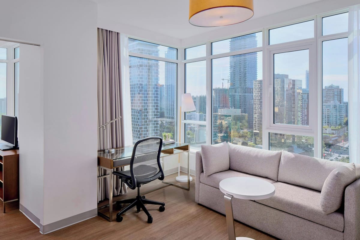 A modern hotel room with a large window, a sofa, a desk with a chair, and a small table.