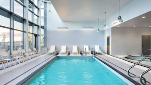 Una piscina interior moderna con agua azul cristalina, tumbonas y grandes ventanales con vistas a la ciudad.